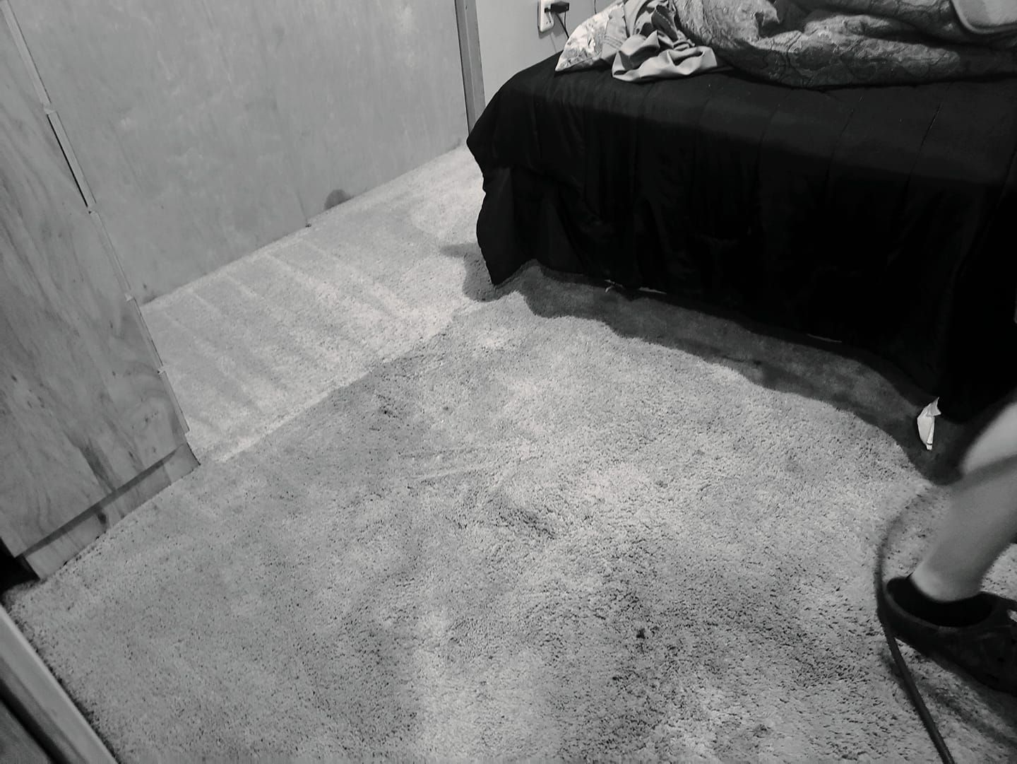 A person stands near a bed in a room with light-colored, textured carpeting.