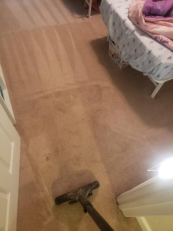 A person uses a carpet cleaning vacuum wand to clean a section of tan carpet in a bedroom near a bed.