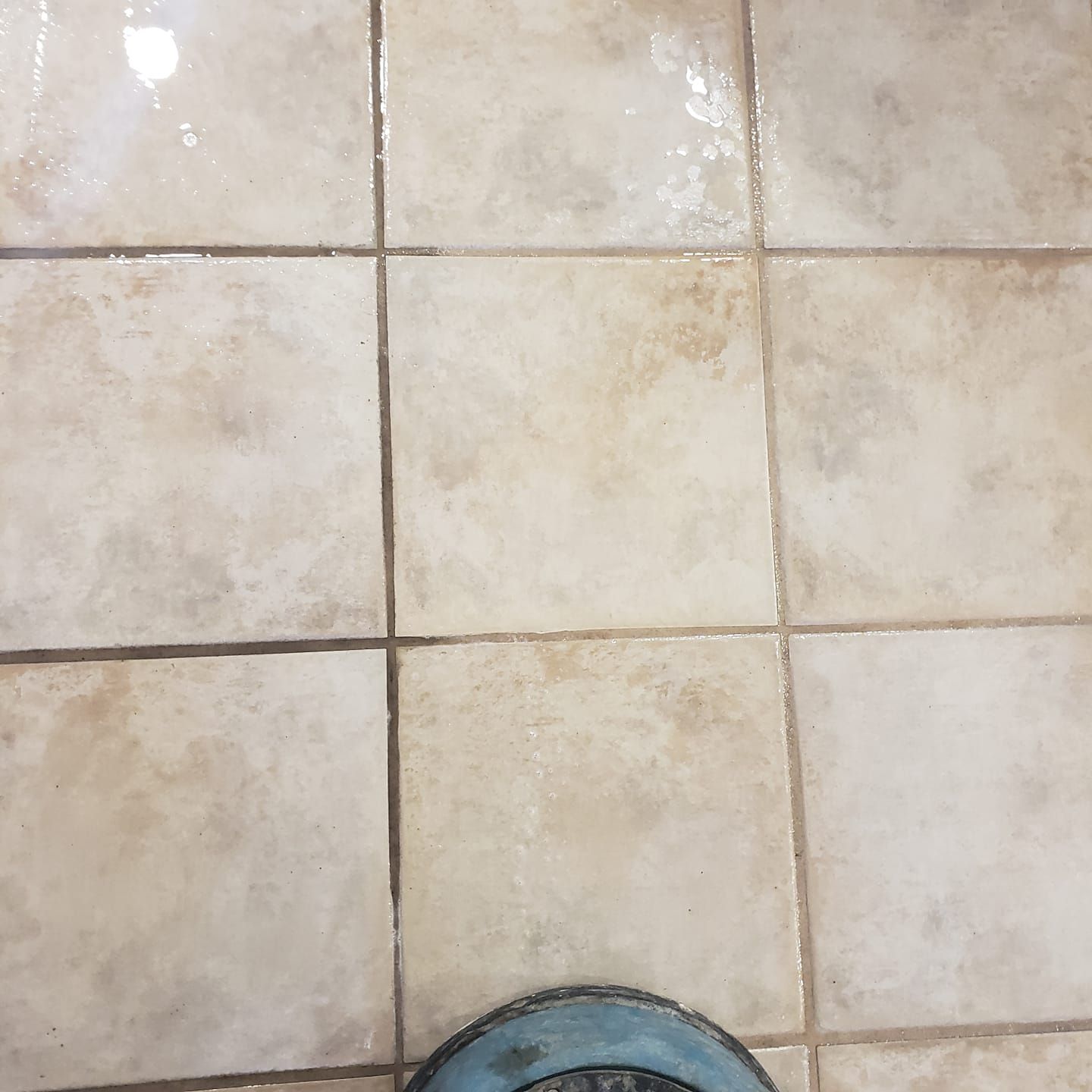 A high-angle view of beige square floor tiles with grout lines, partially obscured by a grey circular object at the bottom.