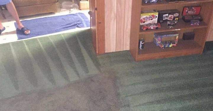 A room with green carpet, a wooden shelving unit with toys, and a person standing near a blue rug.