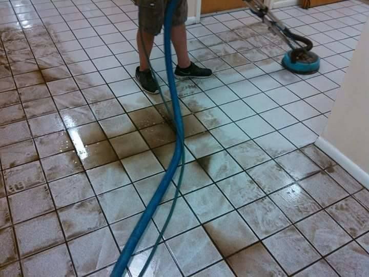 A person uses a circular machine to deep clean dirty white tiled flooring, revealing a clean path behind the tool.