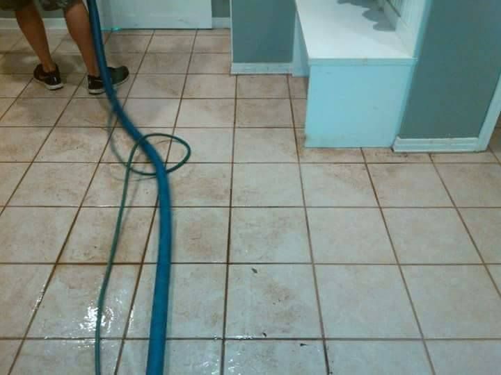 A person uses a professional cleaning hose to clean light-colored, stained tile flooring in a room.