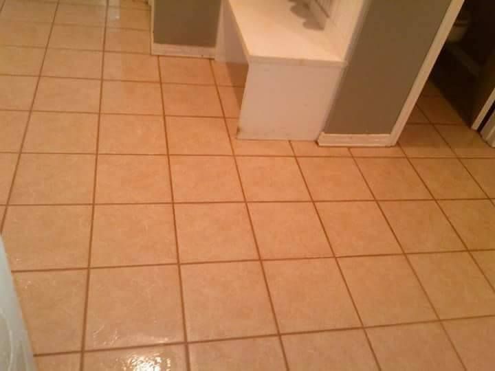 A tiled floor with beige square tiles and grout lines, extending toward a white wall partition in a room.