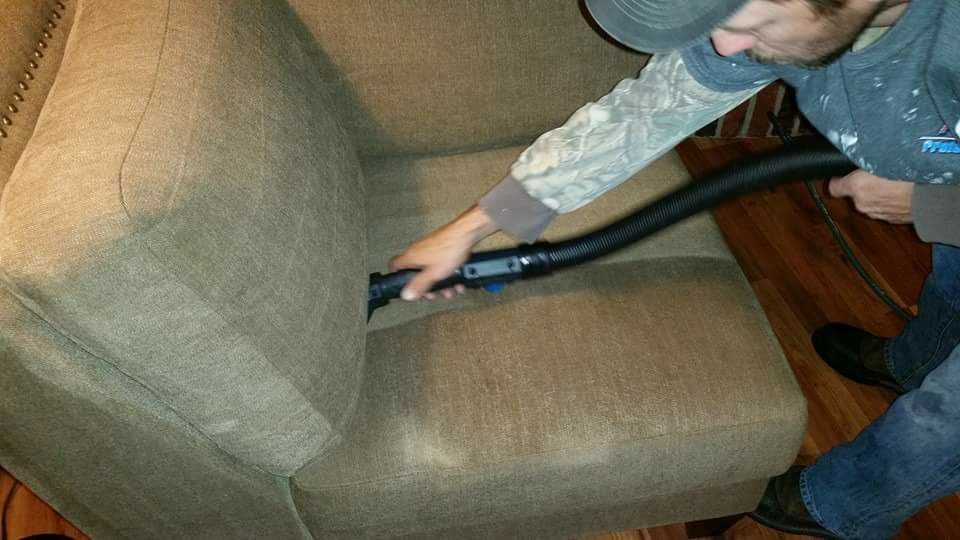 A person vacuuming the crevice of a brown fabric armchair using a black vacuum attachment.