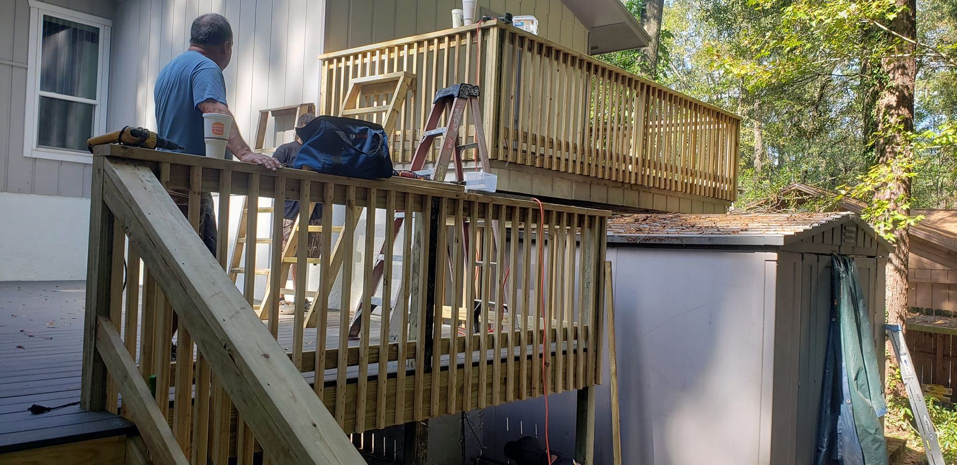 A person stands on a wooden deck next to a house while working on the elevated railing.