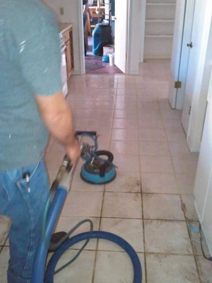 A person cleans light-colored tile flooring using a circular industrial floor scrubber with a connected hose.
