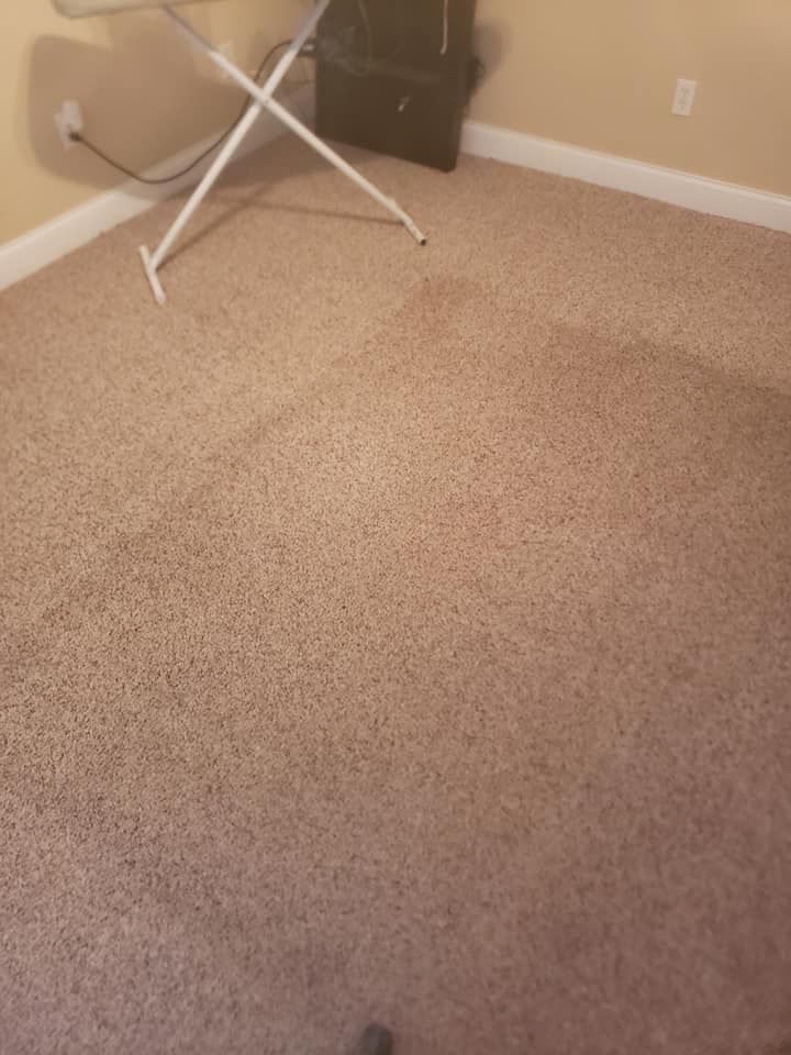 A tan carpeted room showing a square indentation where furniture previously stood, with an ironing board in the corner.