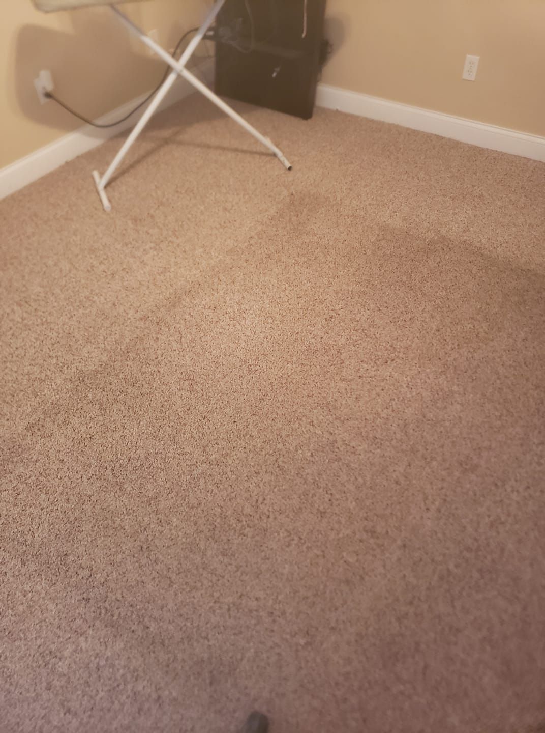 A high-angle view of a beige carpeted floor with an ironing board in the upper left corner and a baseboard along the wall.