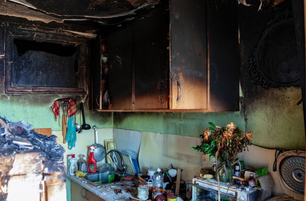 Kitchen after a fire, featuring smoke-damaged walls, charred cabinets, and soot-covered counters and appliances.
