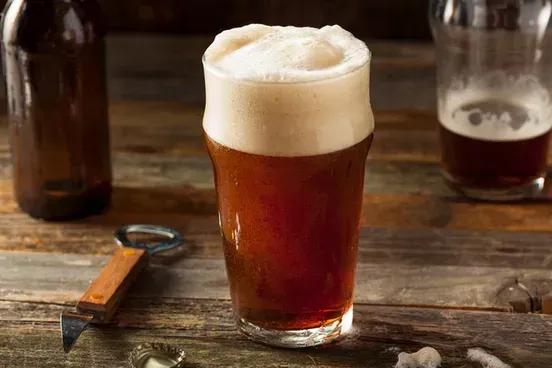 A glass of beer is sitting on a wooden table next to a bottle opener.