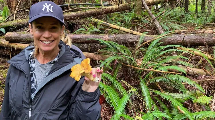 A woman wearing a hat is holding a mushroom in the woods near Woods End Landing in Bamfield BC