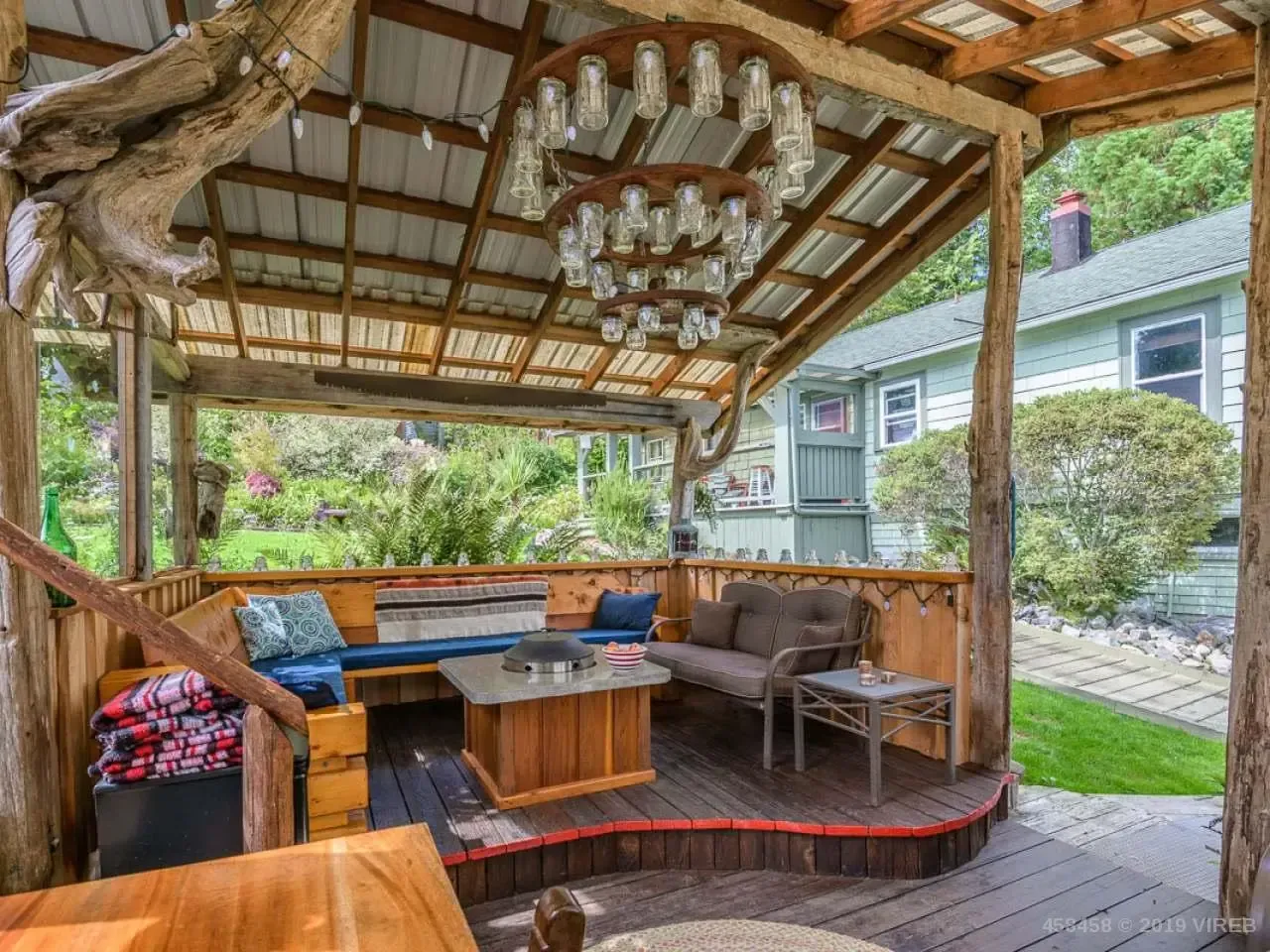 A wooden gazebo with a couch , table and chairs under a roof.