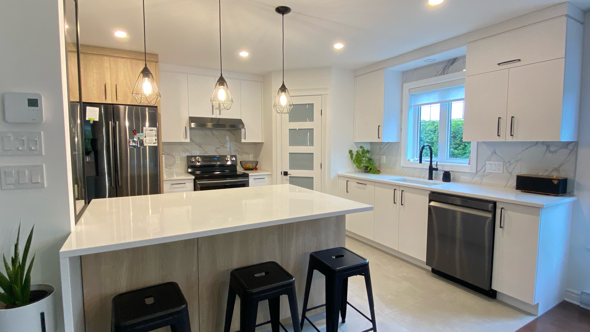 Cuisine blanche moderne avec îlot central, tabourets noirs, appareils électroménagers en acier inoxydable et suspensions lumineuses.