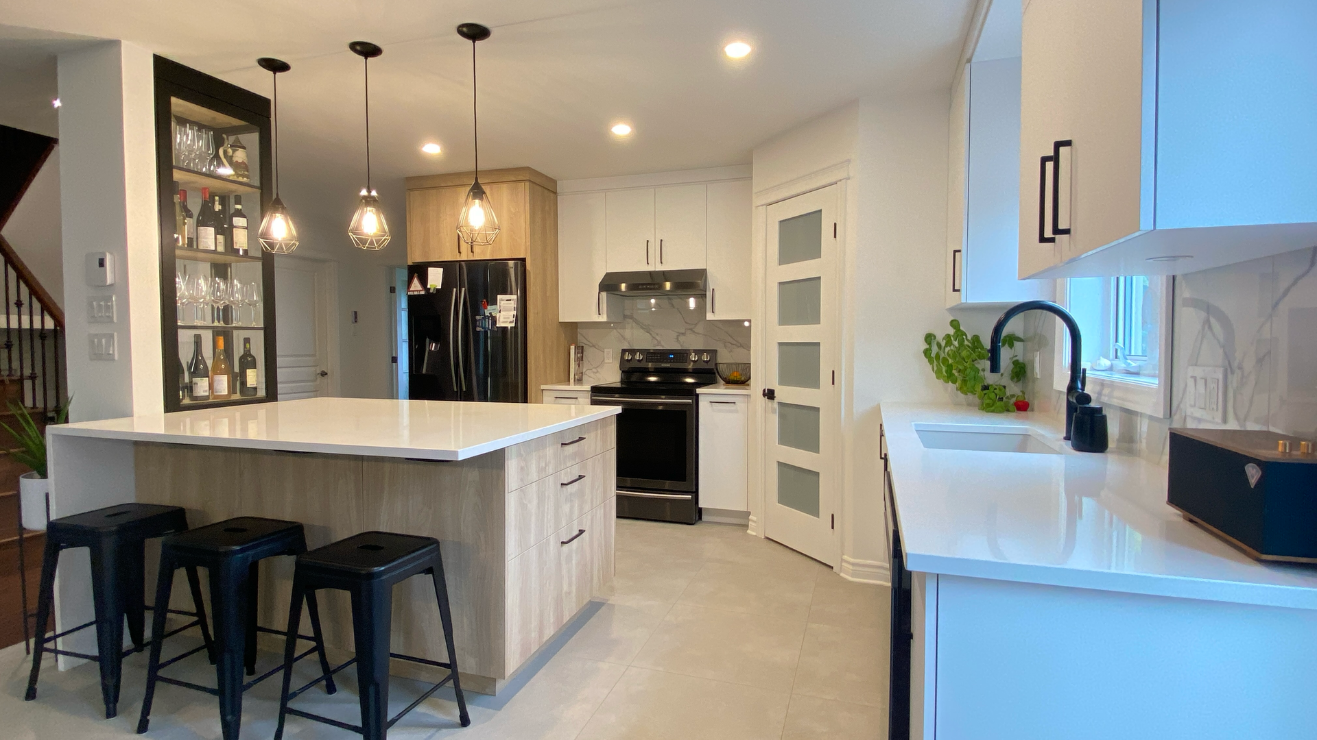 Cuisine moderne avec comptoirs blancs, îlot en bois clair, électroménagers noirs et luminaires suspendus.