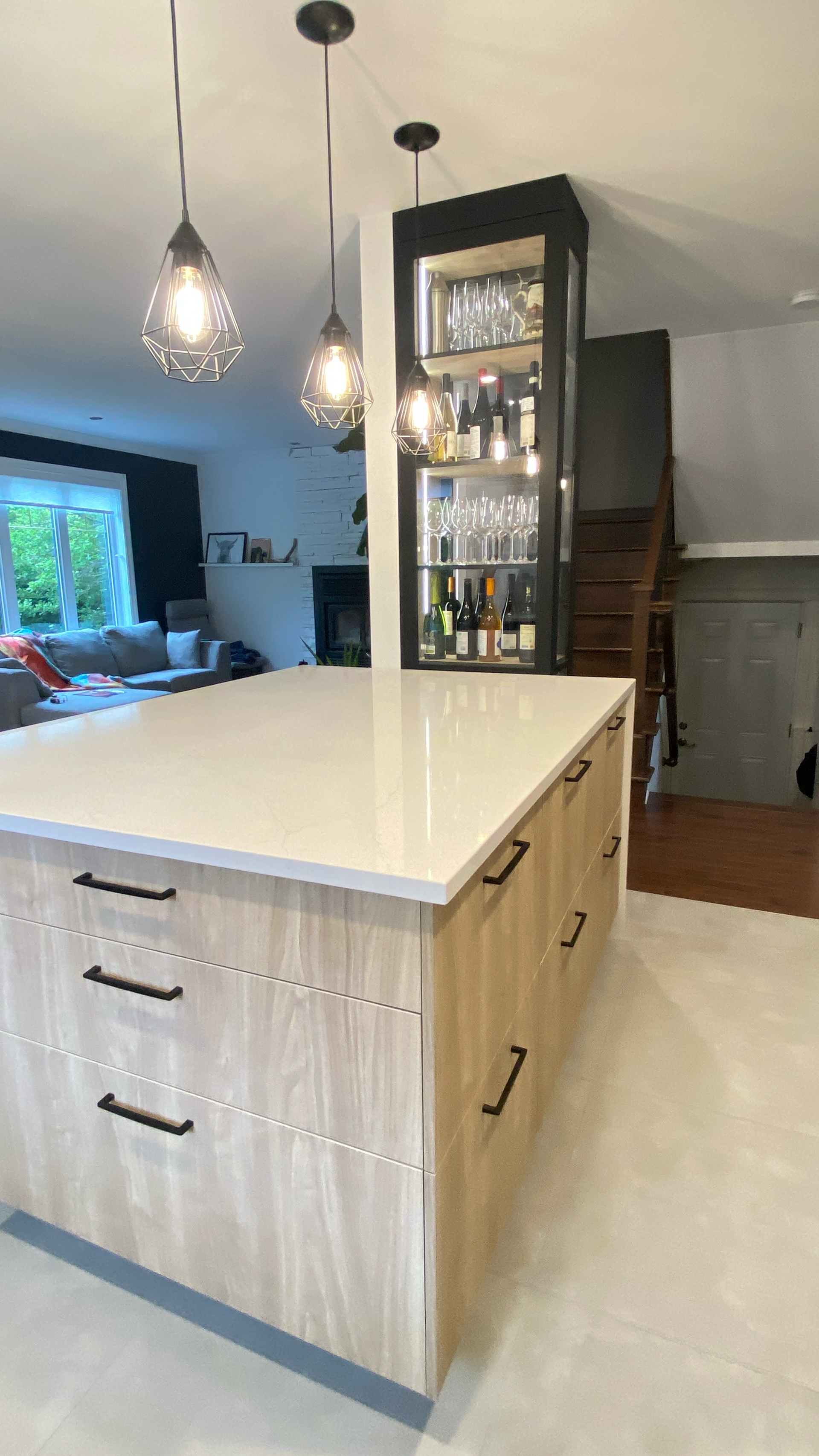 Îlot de cuisine avec placards en bois clair, plan de travail blanc et suspensions lumineuses. Un meuble contenant de la verrerie et des spiritueux est visible à l'arrière-plan.