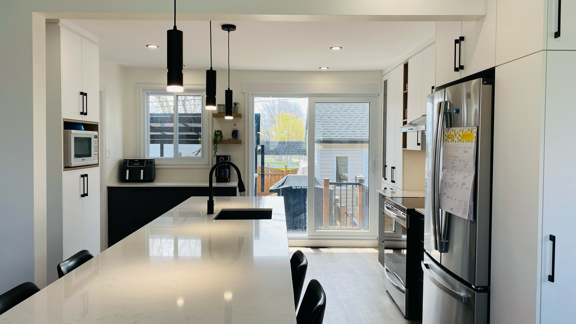 Cuisine moderne avec armoires blanches, îlot central foncé et suspensions lumineuses. Une porte-fenêtre coulissante donne sur le jardin.