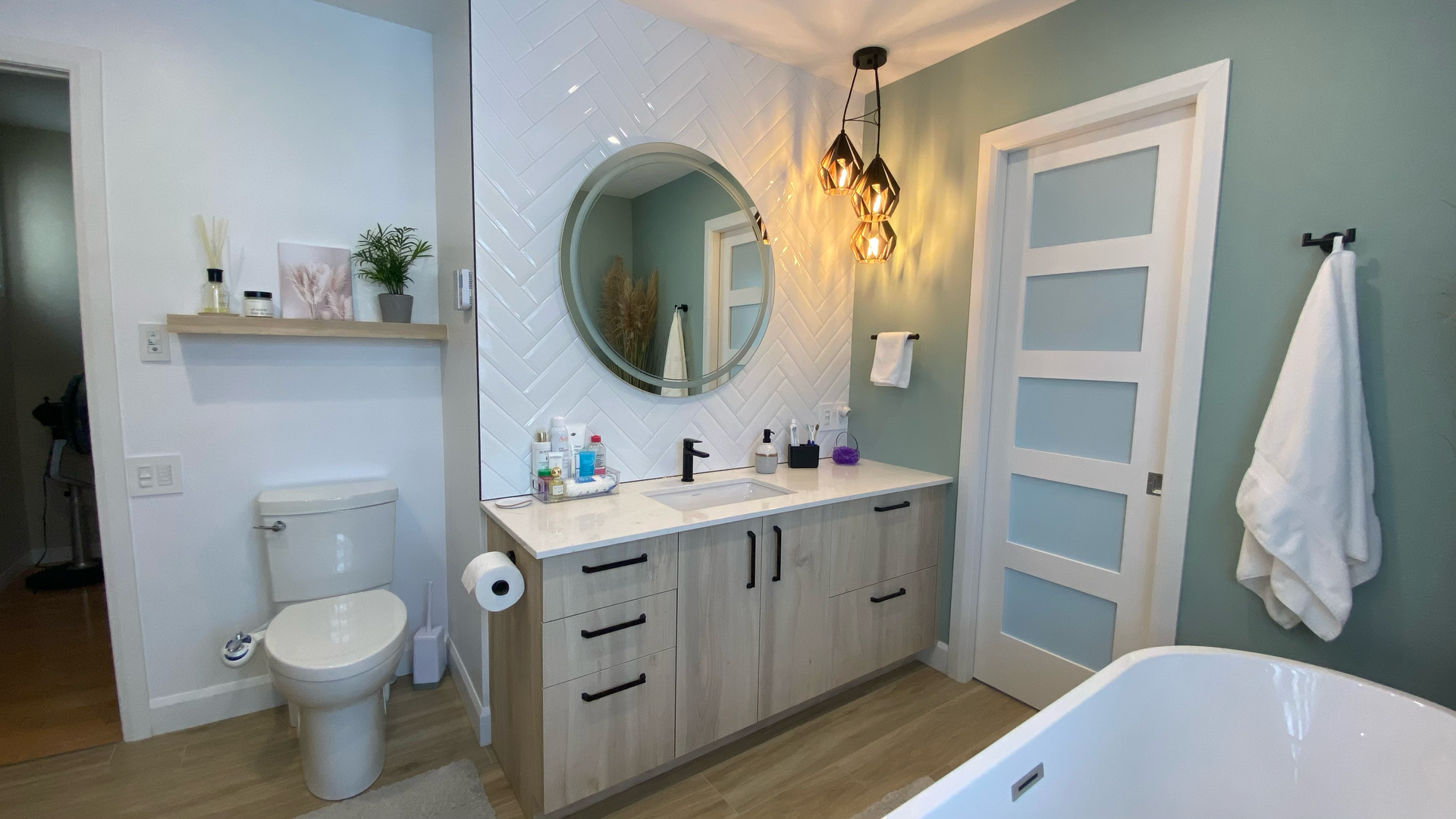 Salle de bains moderne avec meuble-lavabo clair, miroir rond et crédence en carrelage à motifs. Murs vert clair et baignoire blanche.