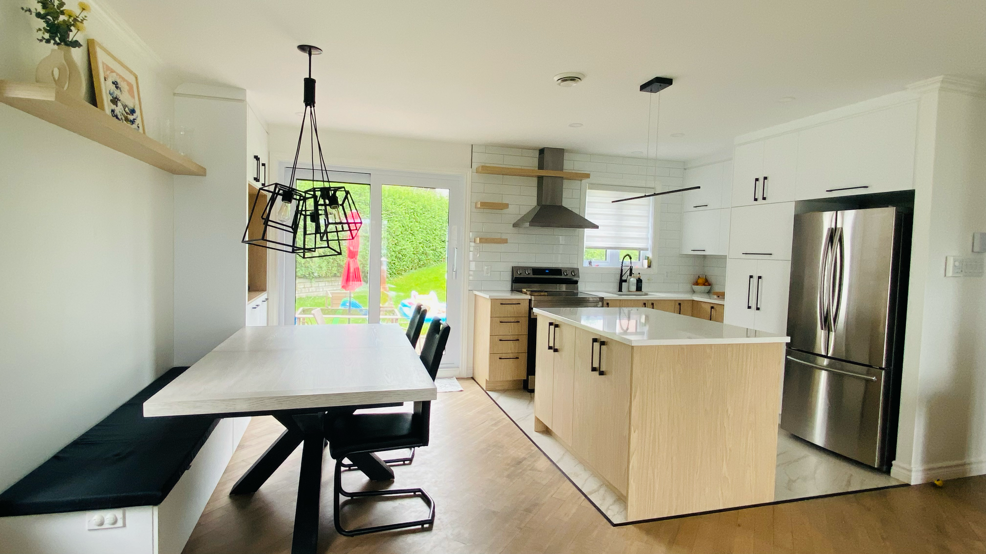 Cuisine moderne avec îlot central et coin repas ; placards en bois clair et blancs, appareils électroménagers en acier inoxydable et grandes fenêtres.