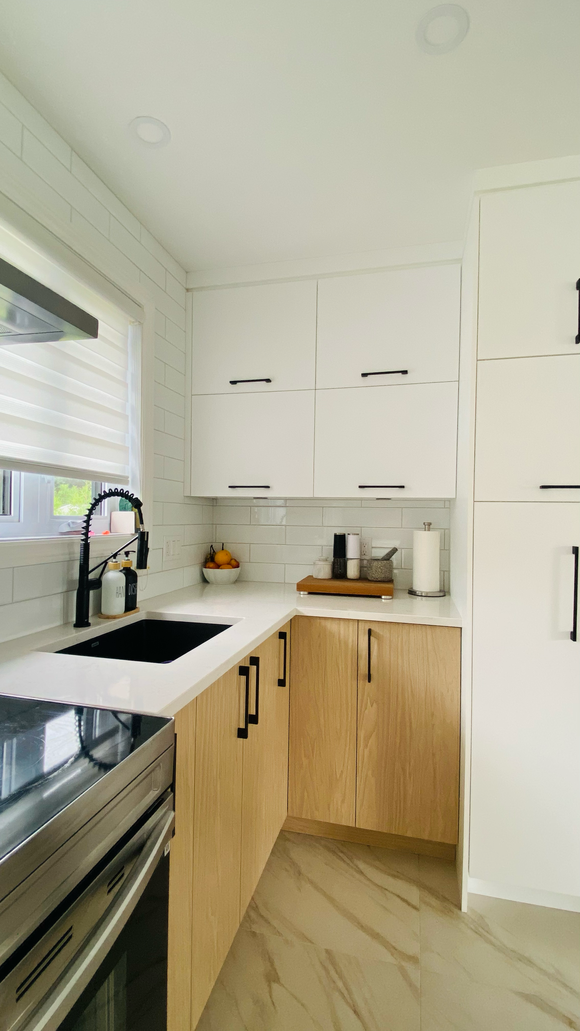 Cuisine moderne avec placards blancs et en bois, évier et robinet noirs, plans de travail blancs, four.
