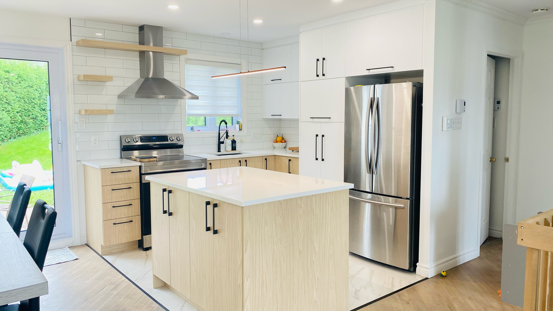 Cuisine moderne avec armoires en bois clair, comptoirs blancs, électroménagers en acier inoxydable et îlot central.