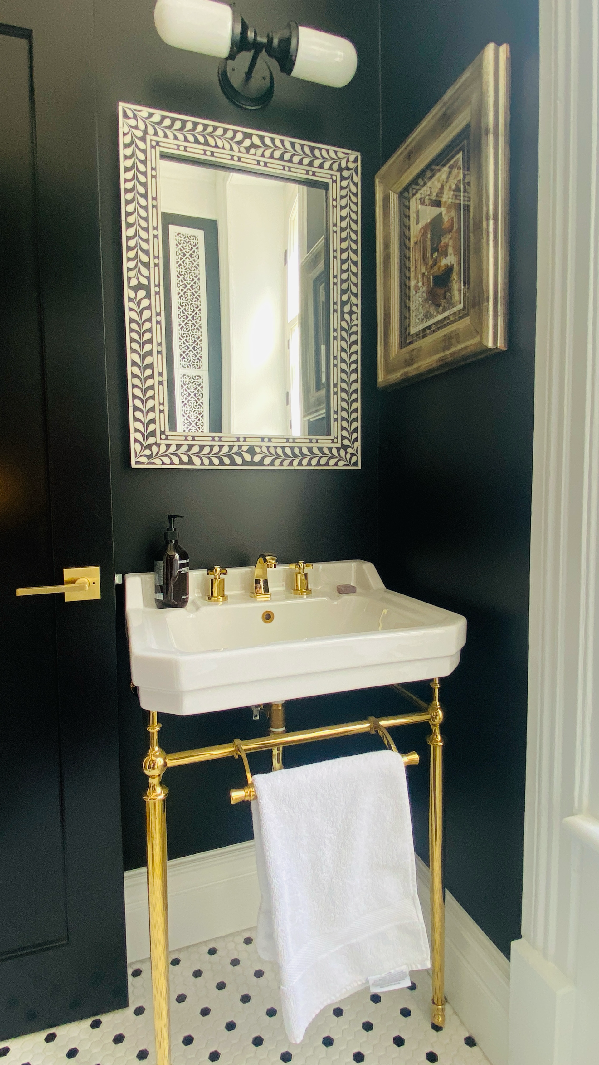 Petite salle de bains aux murs noirs, lavabo blanc avec robinetterie dorée, miroir décoratif et tableau encadré.