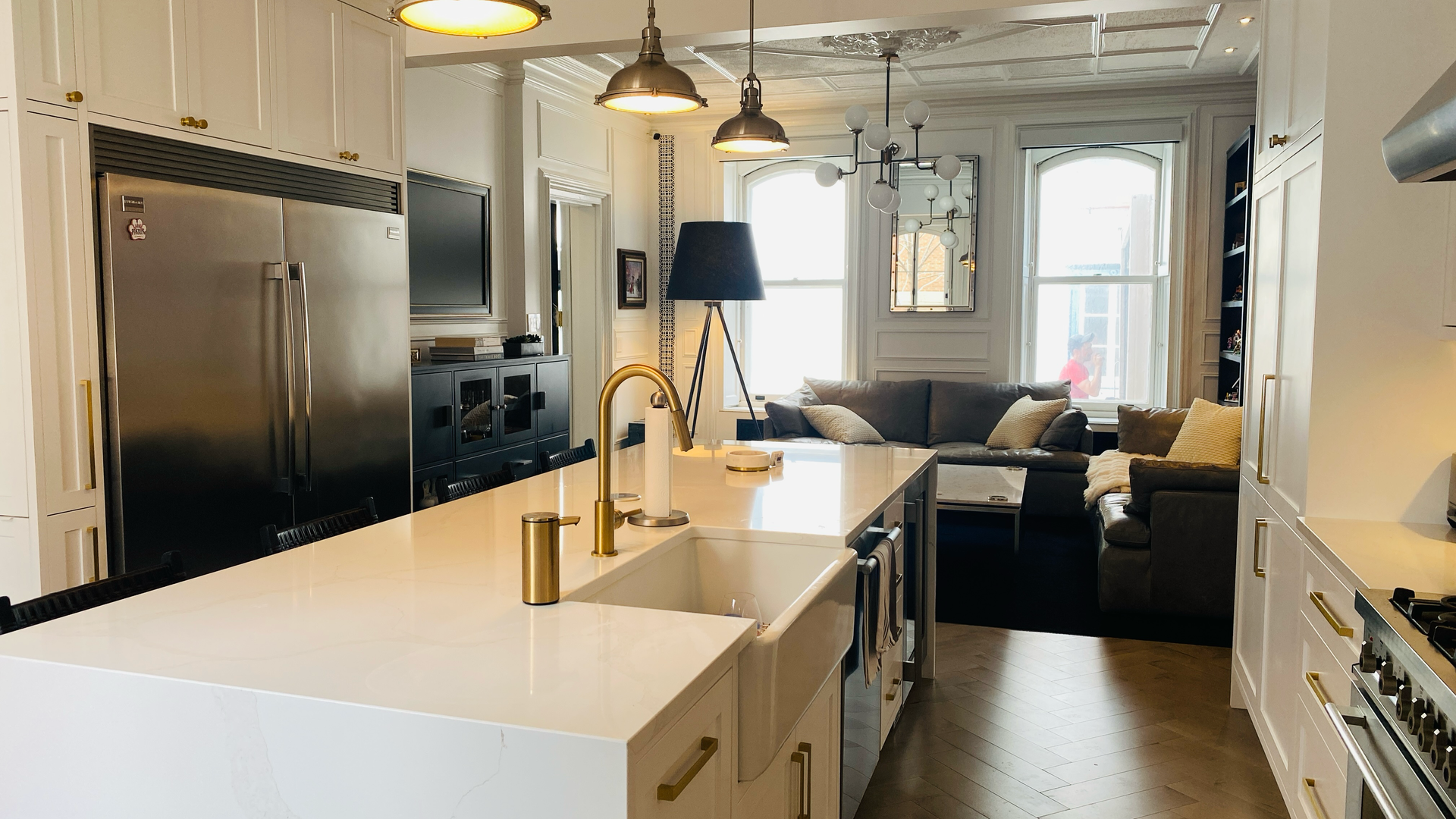 Cuisine moderne avec comptoirs blancs, appareils électroménagers en acier inoxydable et touches dorées, donnant sur un salon avec un canapé gris.