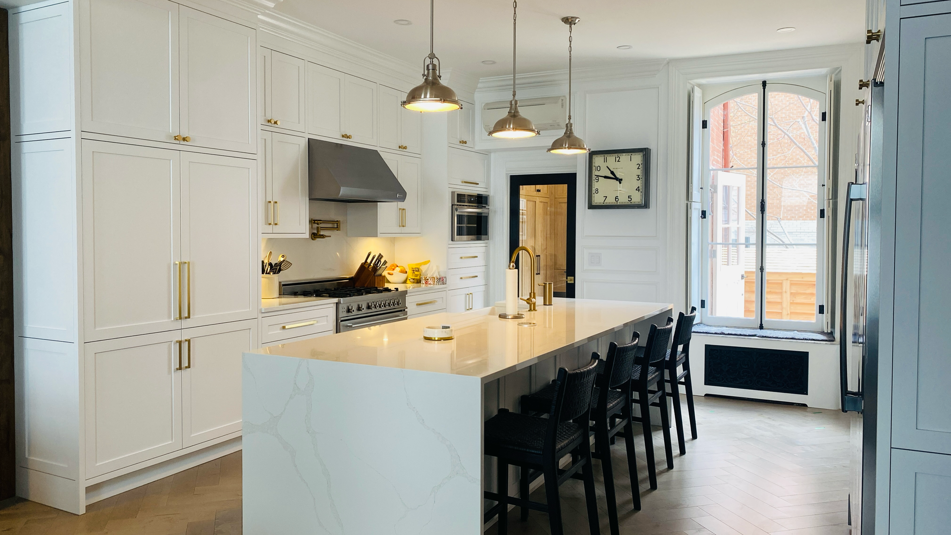 Cuisine blanche avec îlot central, suspensions lumineuses, appareils électroménagers en acier inoxydable, tabourets noirs et grande fenêtre.