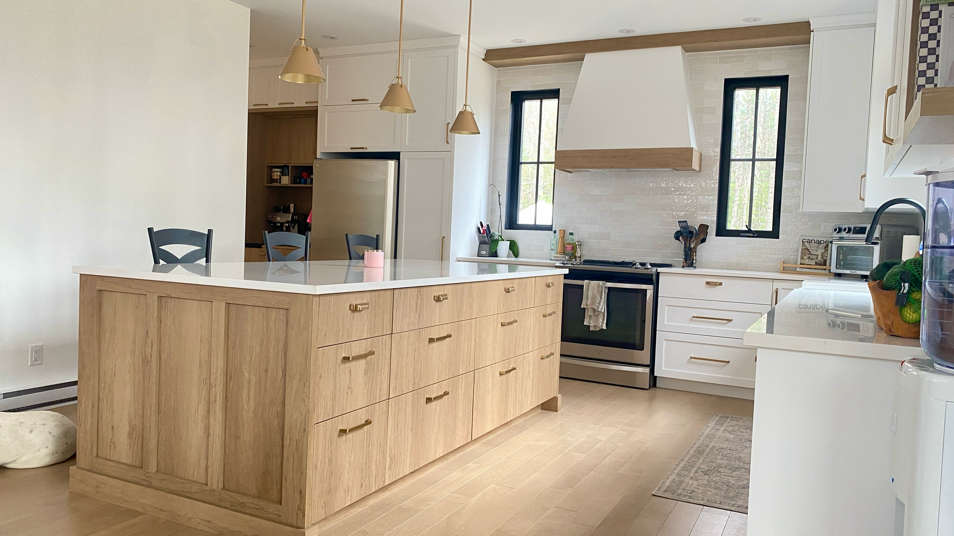 Cuisine spacieuse avec îlot central en bois clair, placards blancs, appareils électroménagers en acier inoxydable et fenêtres à cadre noir.