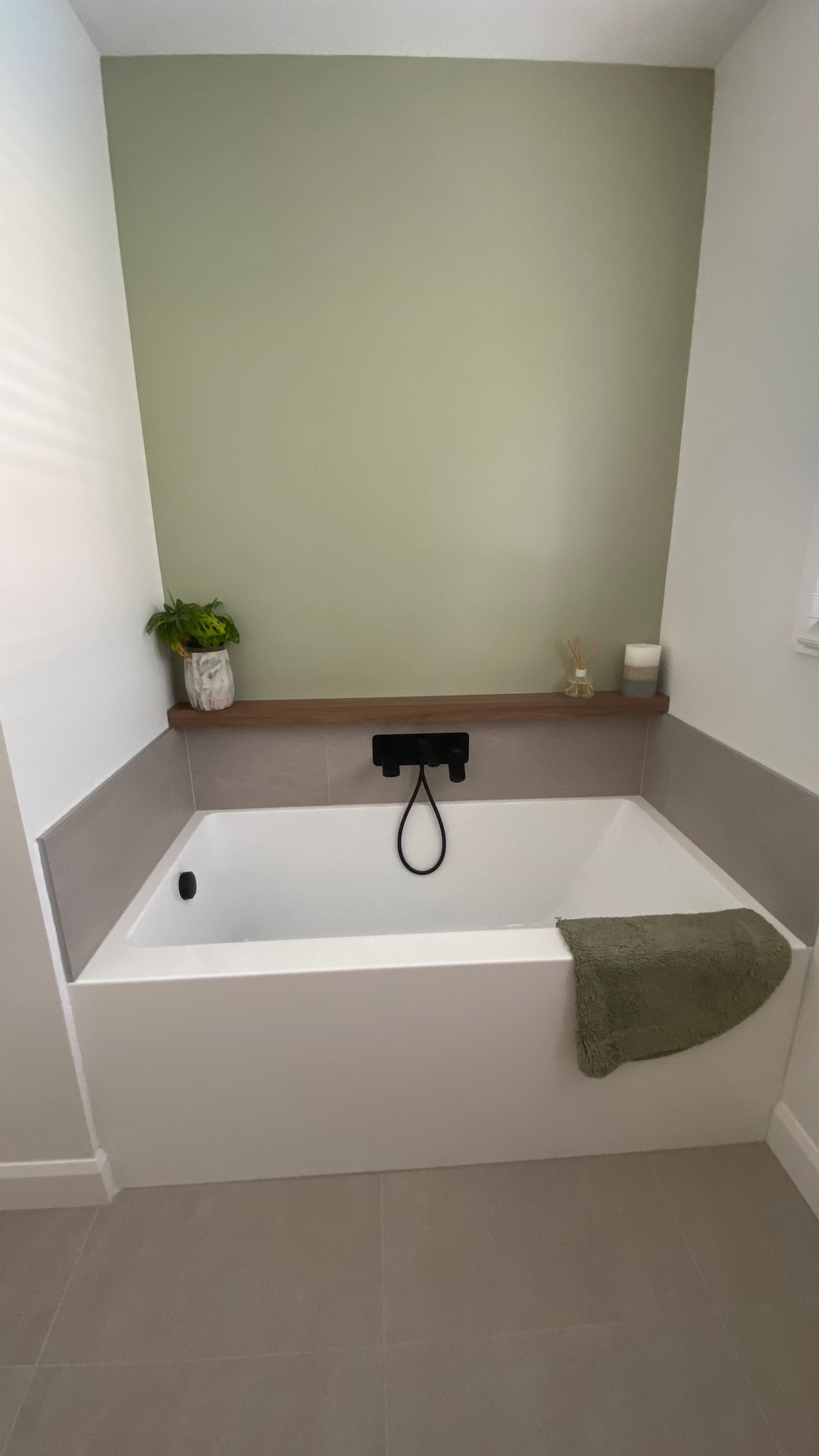 Salle de bains moderne avec baignoire blanche, mur vert, étagère en bois avec plante et bougie.