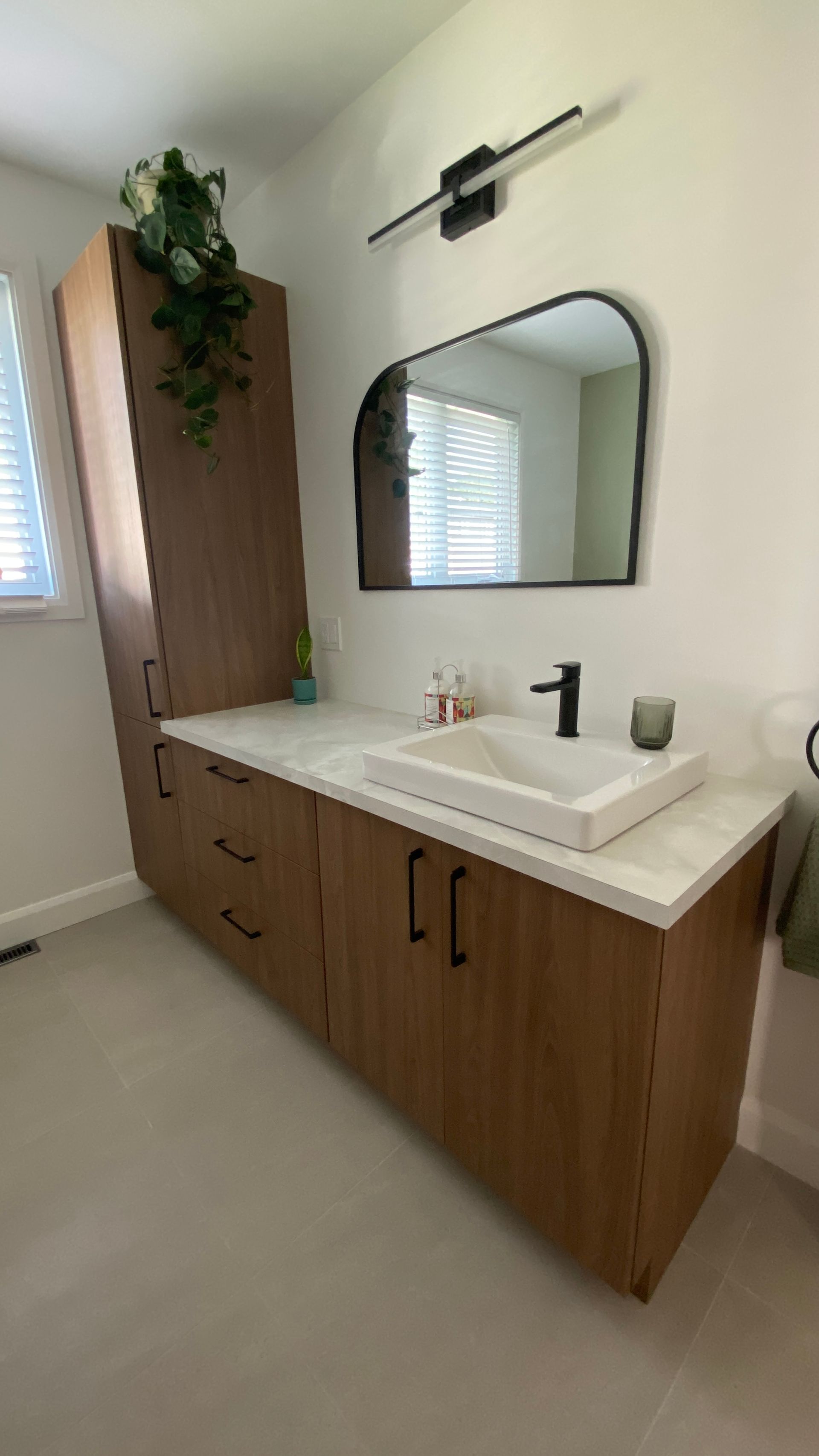 Salle de bains moderne avec meubles en bois, comptoir blanc, robinetterie noire et miroir arqué.
