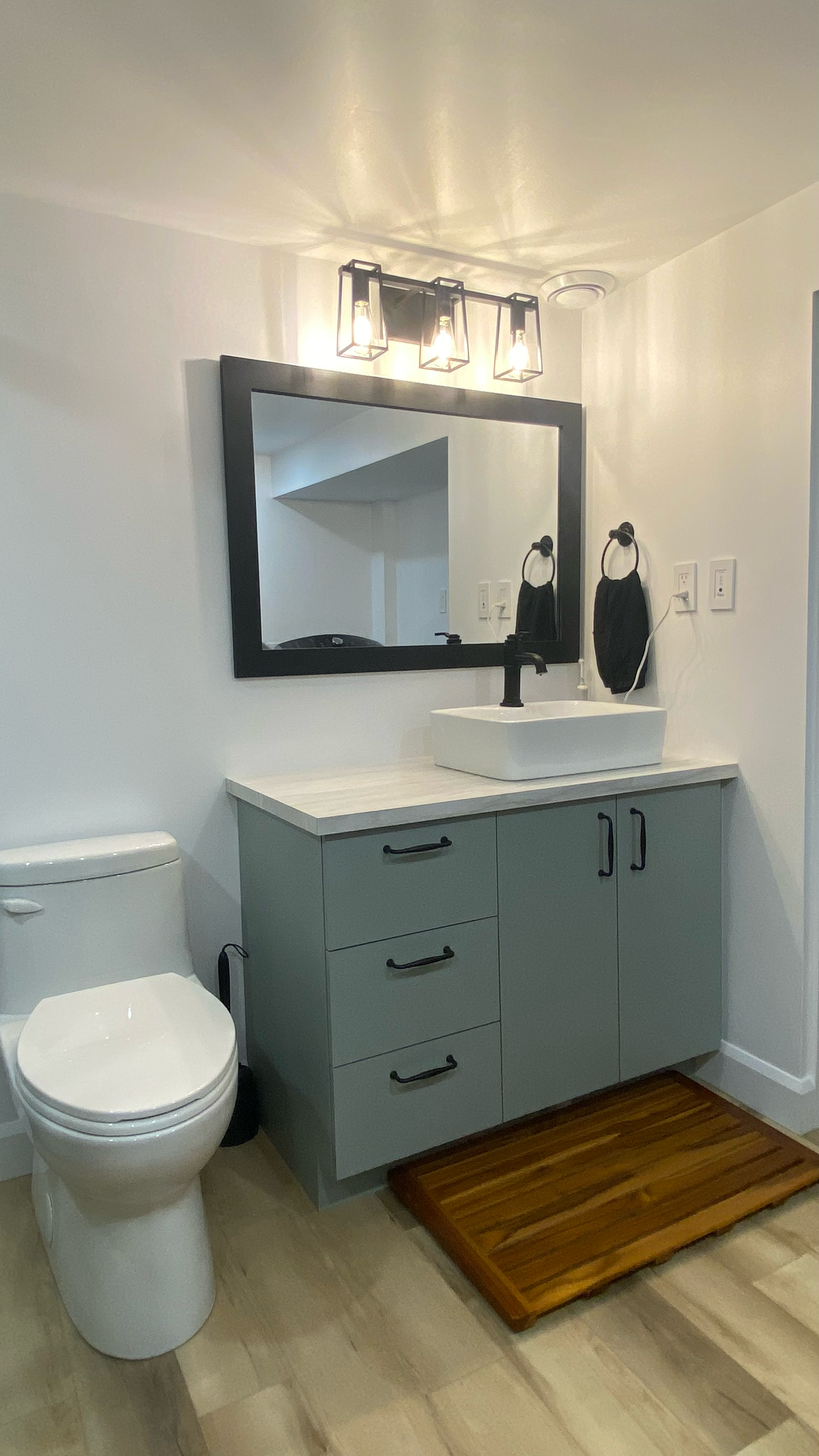 Salle de bains avec meuble-lavabo gris, lavabo blanc, miroir à cadre noir et toilettes. Un tapis en bois est posé au sol.