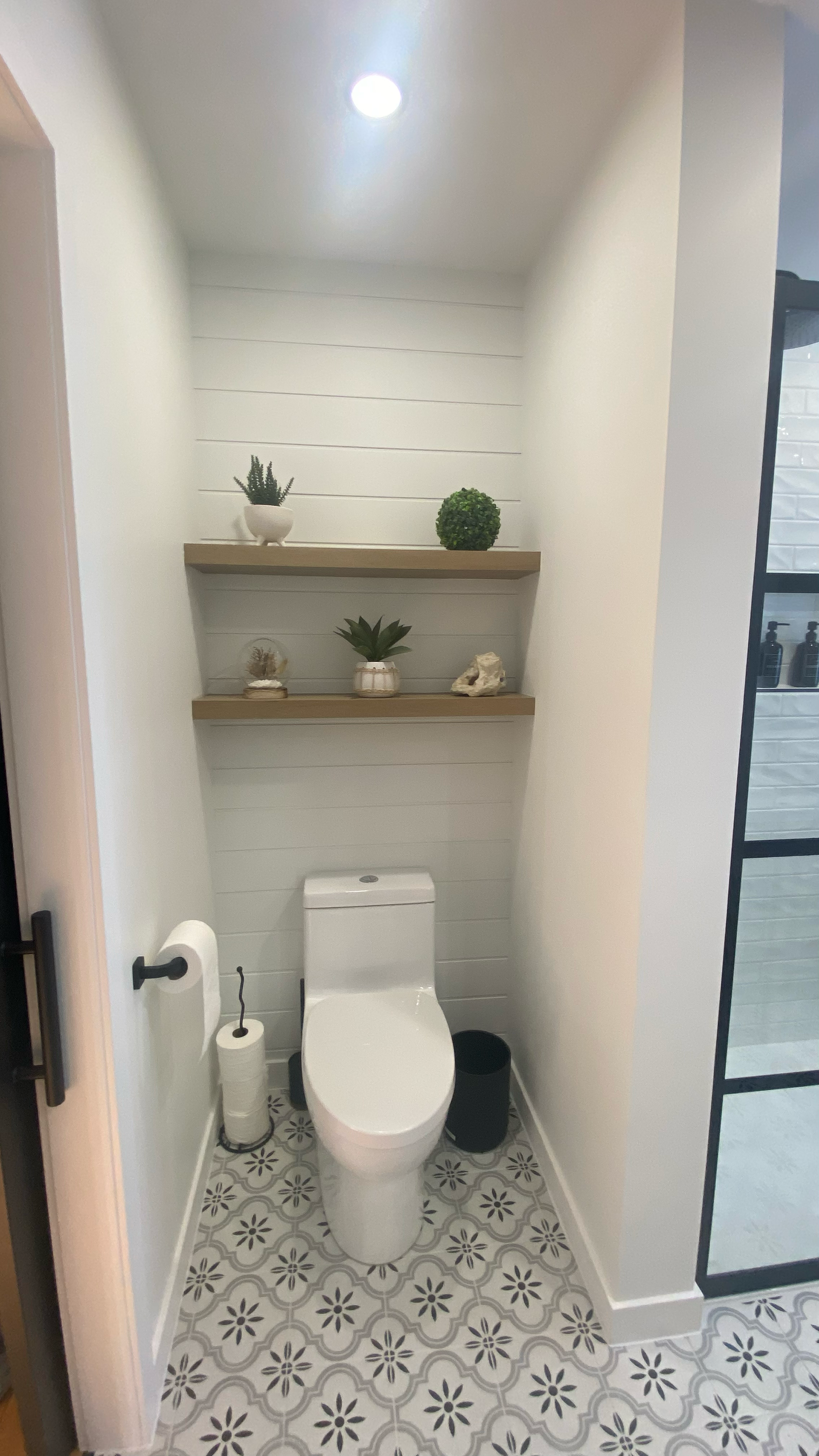 Petite salle de bains avec sol à motifs, toilettes blanches, étagères avec plantes et mur carrelé de blanc texturé.
