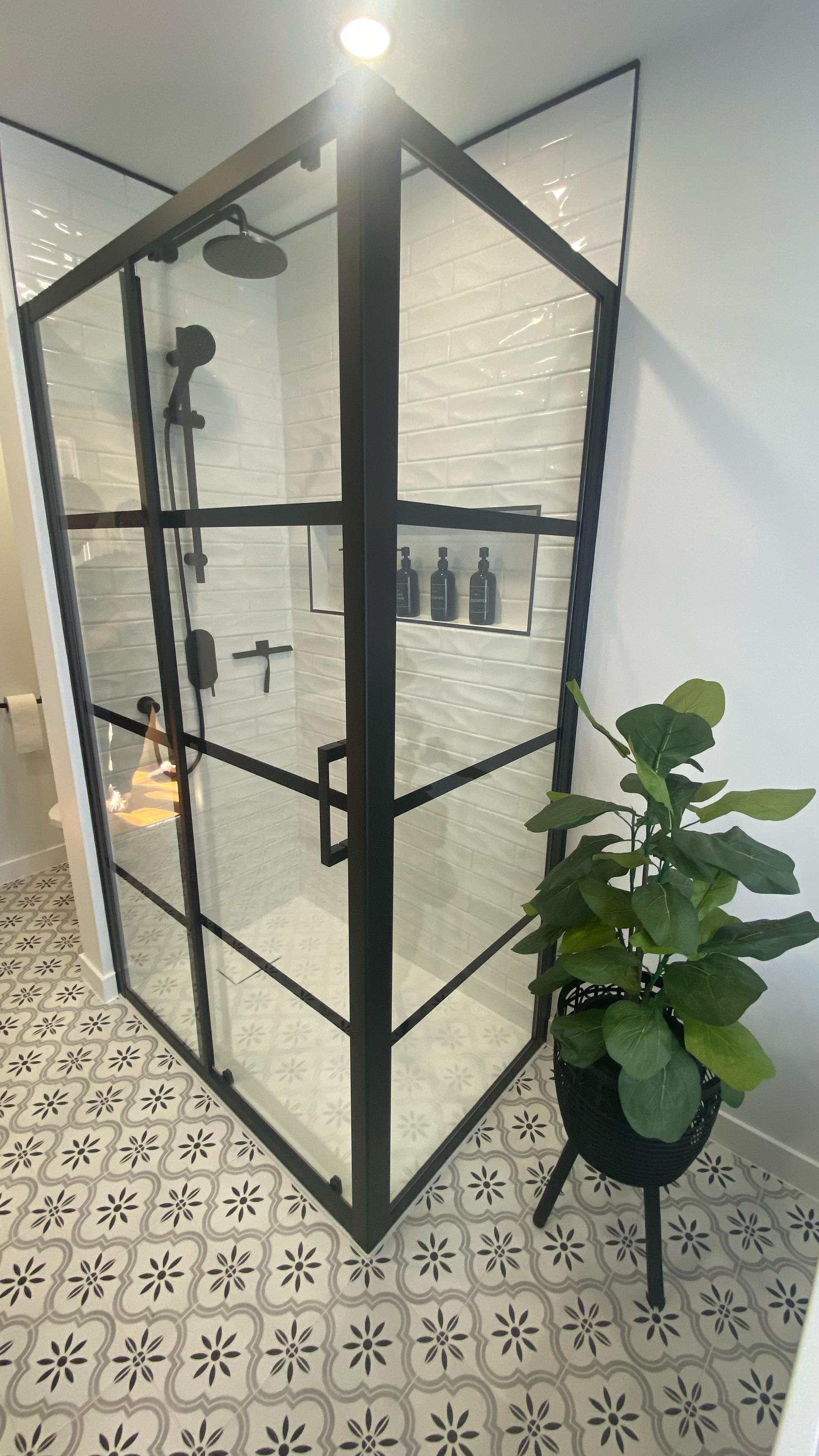 Douche en verre à cadre noir dans une salle de bains avec sol carrelé à motifs, mur carrelé de blanc texturé et plante verte en pot.