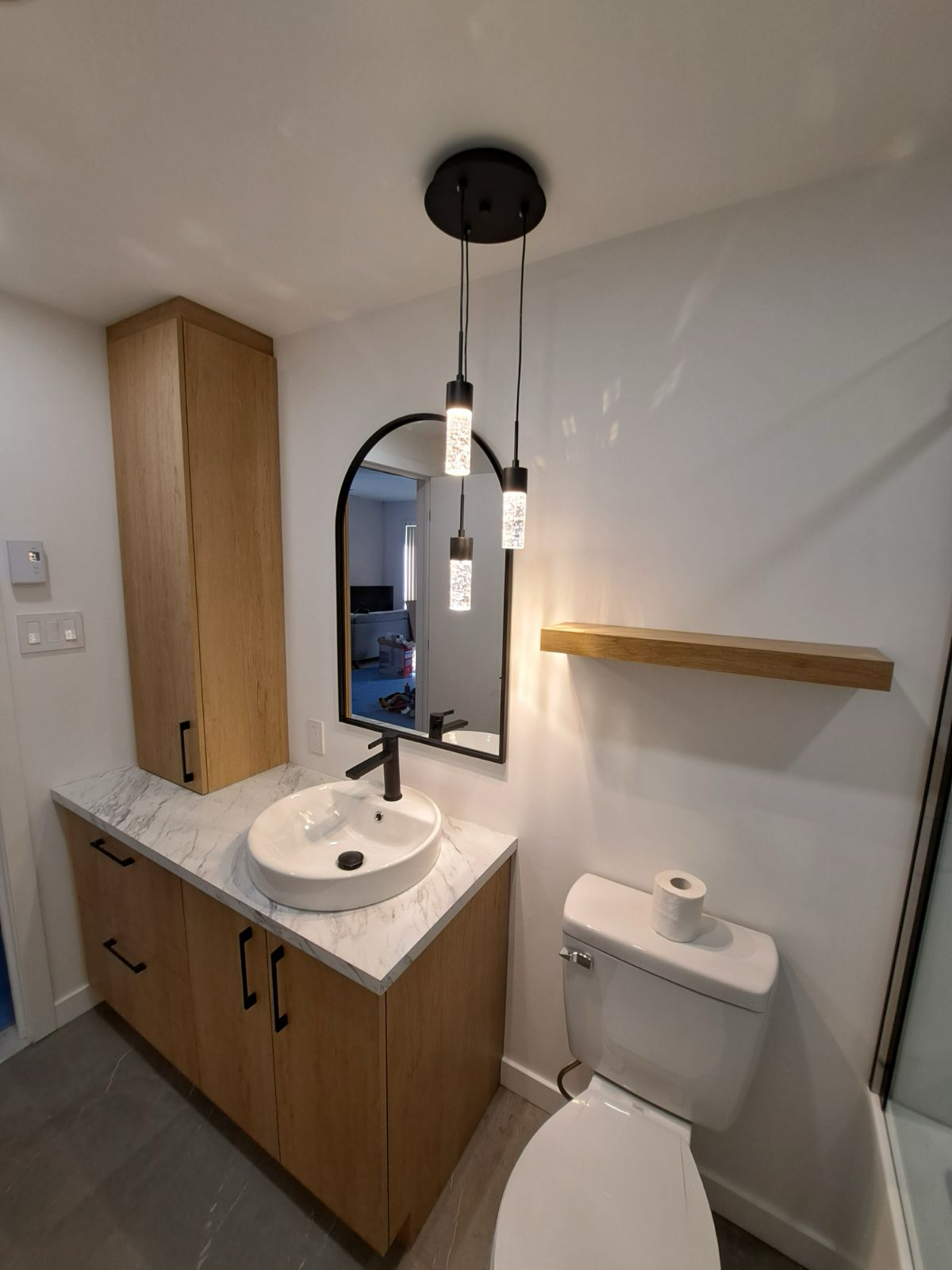 Salle de bains moderne avec meuble-lavabo en chêne, miroir arqué noir, suspensions lumineuses et toilettes.