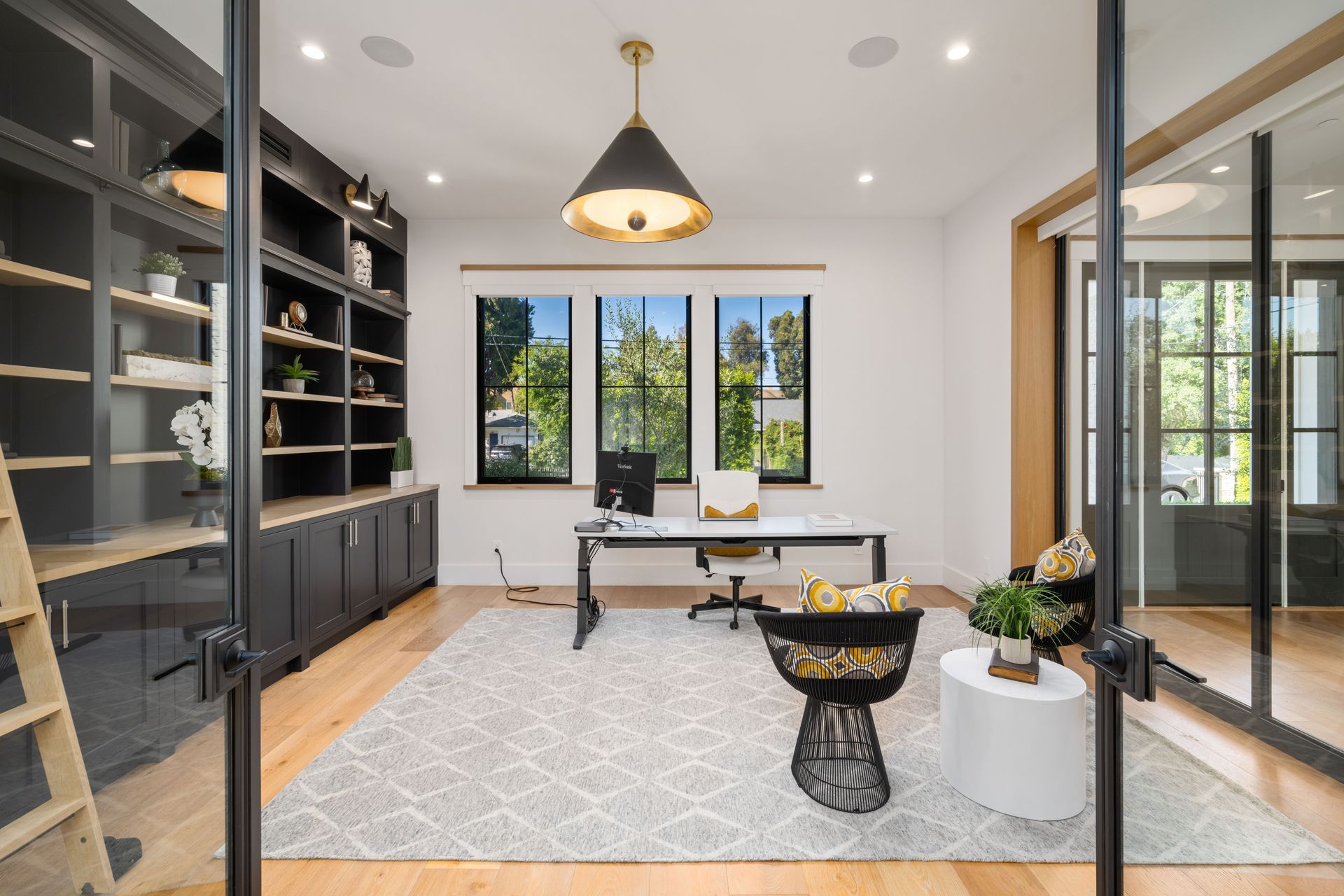 Bureau avec étagères noires, bureau et grandes fenêtres. Comprend un tapis, une chaise et une table décorative.