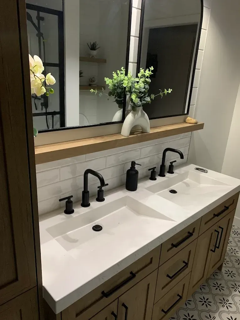 Meuble-lavabo de salle de bain avec deux vasques, robinetterie noire, armoire en bois clair et sol en carrelage décoratif.