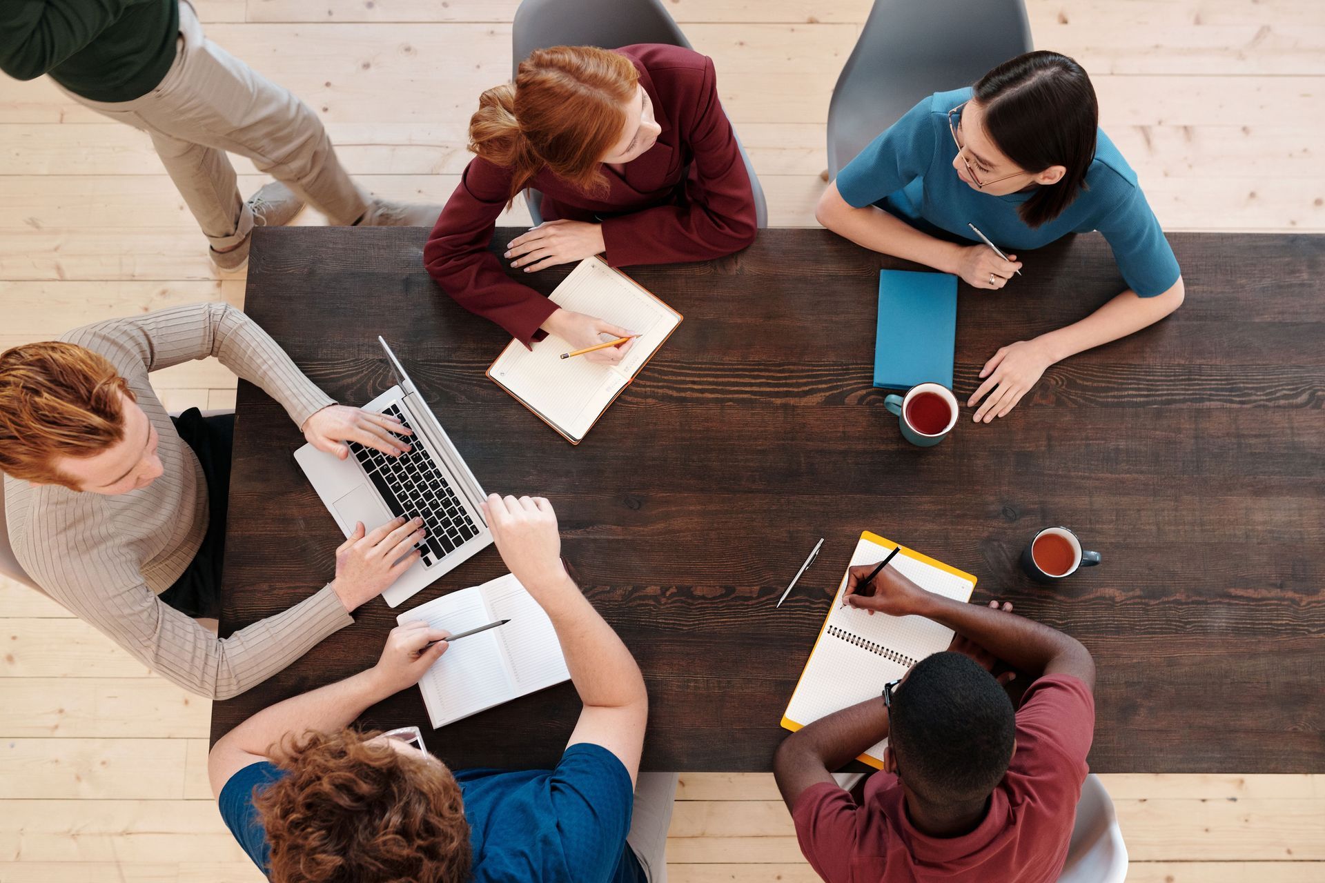 Team meeting around a wooden table, with laptops, notebooks, and coffee mugs from an overhead view