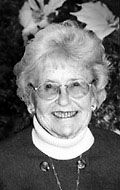 A black-and-white portrait of a smiling person wearing glasses and a high-neck sweater in front of floral decor.