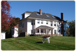 A large, white, multi-story house with a grey roof and brick chimneys stands on a grassy lawn under a clear blue sky.