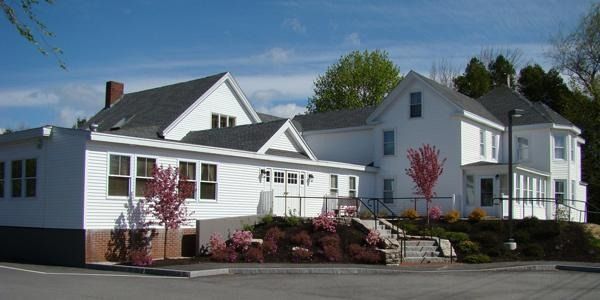 A large, multi-story white building with gabled roofs and a long, single-story wing, set against a bright blue sky.