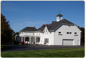 A white farmhouse with a large barn-style garage, a cupola, and a covered porch under a clear blue sky.