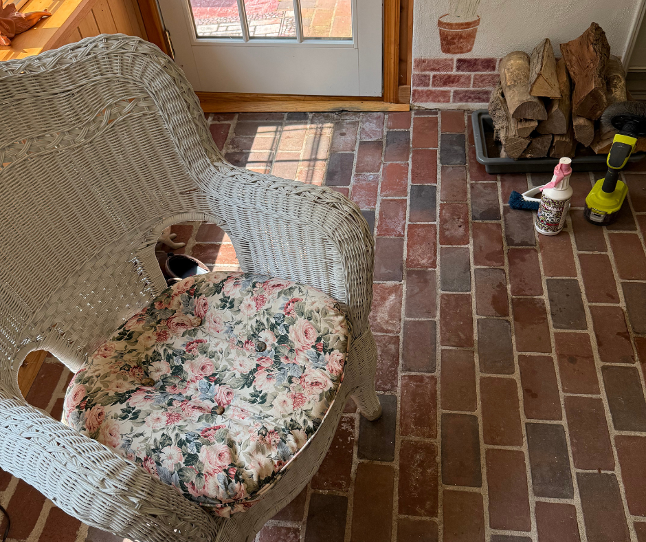 Wicker chair with floral cushion on brick porch floor beside stacked firewood and a doormat