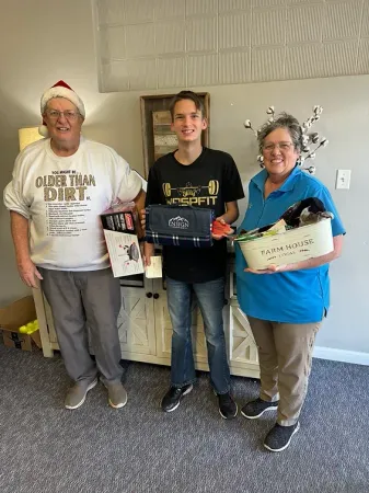 Three people are standing next to each other in a living room holding gifts.