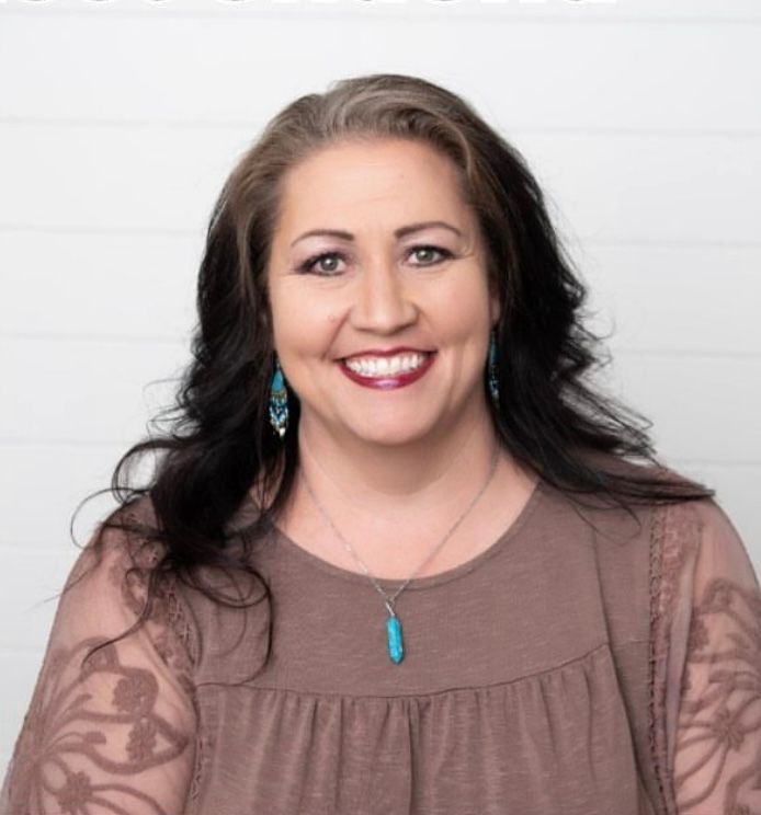 A woman in a blue shirt is smiling in front of a brick wall.