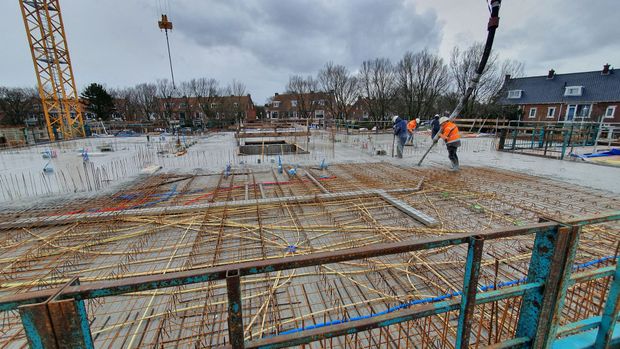 Bouwplaats met wapeningsstaal, arbeiders, kraan en gebouwen op de achtergrond onder een bewolkte hemel.