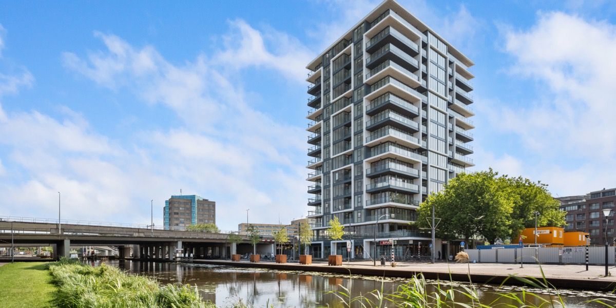 Modern hoogbouwappartementencomplex aan een kanaal onder een blauwe hemel met wolken.