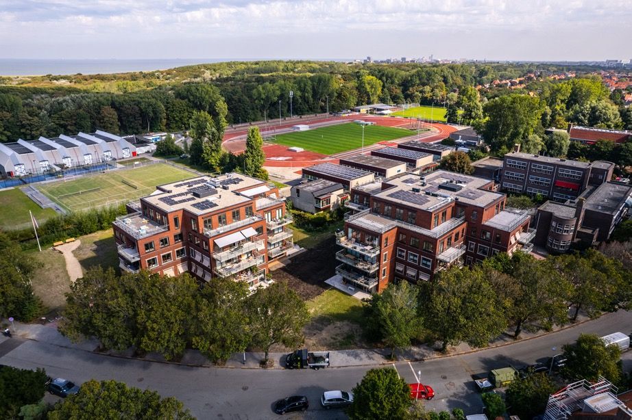 Luchtfoto van bakstenen appartementencomplexen nabij een sportveld en een rij bomen onder een blauwe hemel.