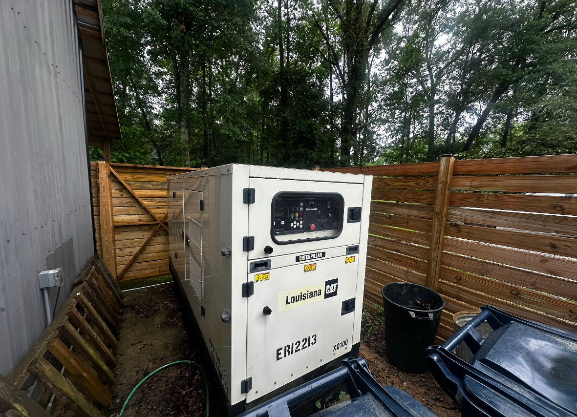White industrial generator in a fenced outdoor yard with a blue truck bed beside it.