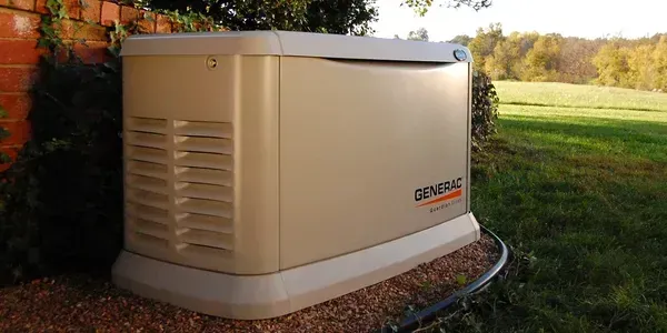 Beige backup generator outdoors beside a house, on a lawn by trees.