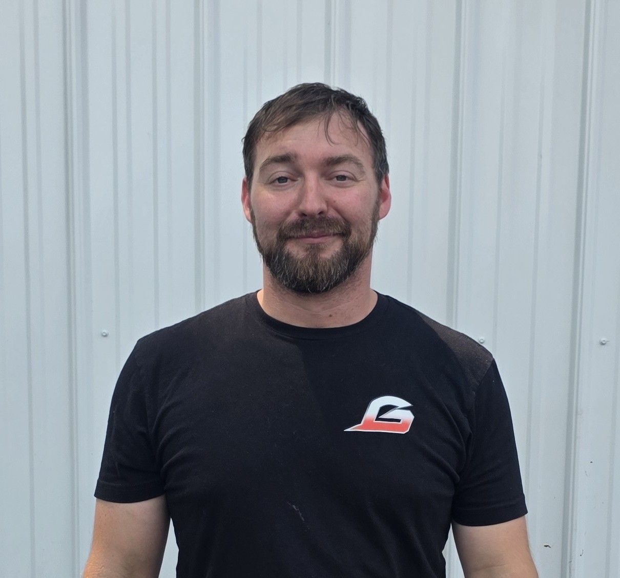 Man in black T-shirt standing against a light corrugated metal wall, smiling at the camera